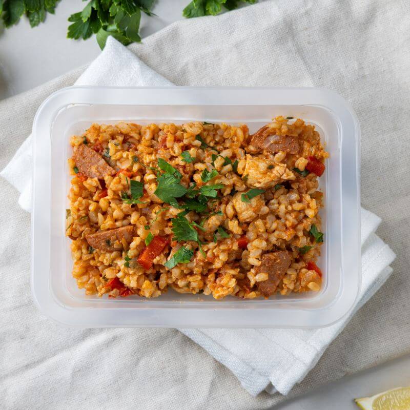 A clear plastic container of Paella with Brown Rice resting on a white napkin and beige cloth. The dish showcases tender chicken, smoky chorizo, and a mix of colorful vegetables atop brown rice, with fresh cilantro in the background adding a vibrant touch.