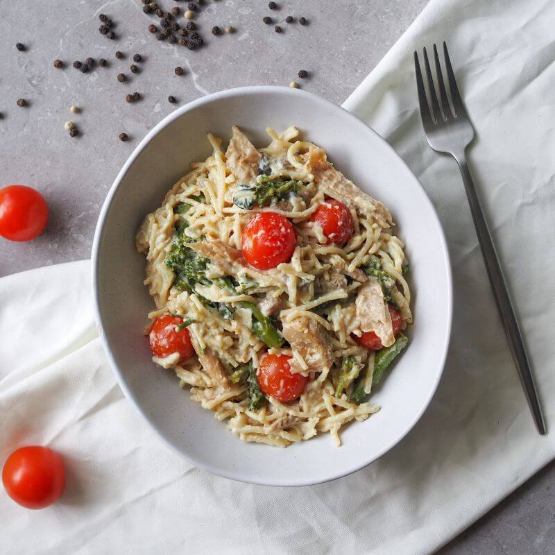 A top-down view of Chicken Alfredo served in a white bowl. Gluten-free spaghetti is coated in a creamy cauliflower and coconut cream Alfredo sauce, paired with tender chicken, pan-roasted cherry tomatoes, broccolini, and mushrooms. The dish is topped with a sprinkle of parmesan. A fork rests to the right of the bowl, with scattered cherry tomatoes and black peppercorns on a grey marble benchtop. A white cloth is tucked underneath the bowl.