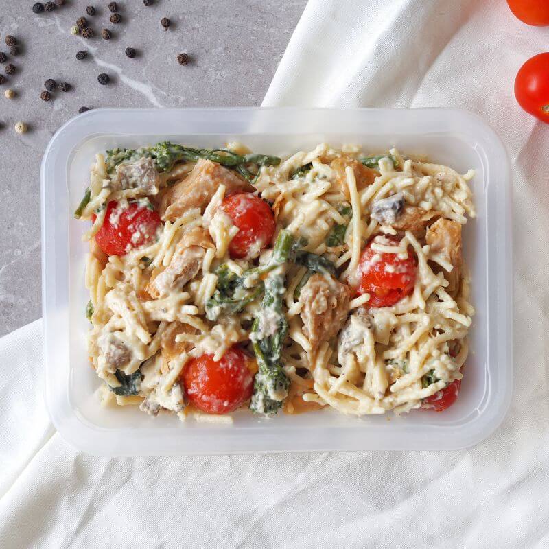 A clear container filled with Chicken Alfredo, featuring gluten-free spaghetti in a creamy cauliflower and coconut cream Alfredo sauce, tender chicken, roasted cherry tomatoes, broccolini, mushrooms, and parmesan. Scattered cherry tomatoes and black peppercorns are visible on the grey marble benchtop. A white cloth is tucked underneath the container.