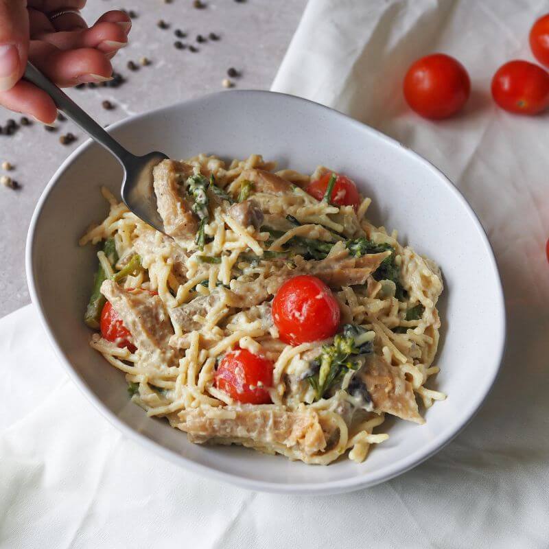 An action shot of Chicken Alfredo. A hand is holding a fork, lifting a piece of tender chicken from the dish. The gluten-free spaghetti is coated in a creamy Alfredo sauce and paired with roasted cherry tomatoes, broccolini, mushrooms, and a sprinkle of parmesan. Scattered cherry tomatoes, black peppercorns, and a white cloth can be seen in the background on a grey marble benchtop.