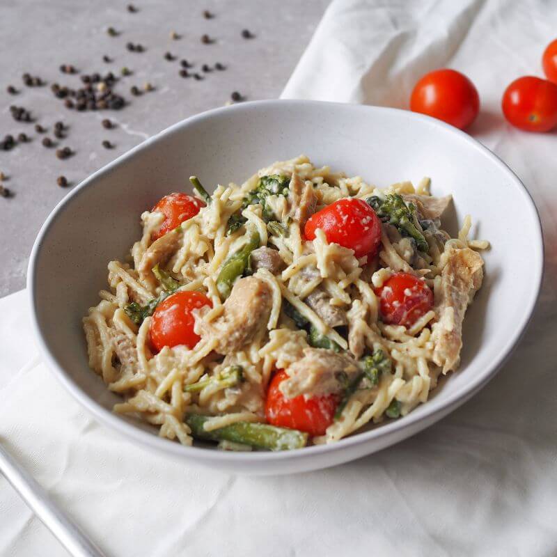 A side angle view of Chicken Alfredo served in a white bowl. The dish features gluten-free spaghetti in a creamy cauliflower and coconut cream Alfredo sauce, paired with succulent chicken, roasted cherry tomatoes, broccolini, mushrooms, and topped with parmesan. Scattered cherry tomatoes and black peppercorns are visible on the grey marble benchtop, with a white cloth tucked underneath the bowl.