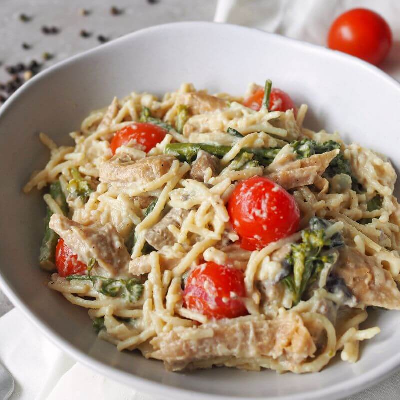 A close-up view of Chicken Alfredo, showcasing gluten-free spaghetti coated in a creamy Alfredo sauce, tender pieces of chicken, roasted cherry tomatoes, broccolini, mushrooms, and a sprinkle of parmesan. The texture of the ingredients is highlighted, with hints of cherry tomatoes and peppercorns visible in the background.