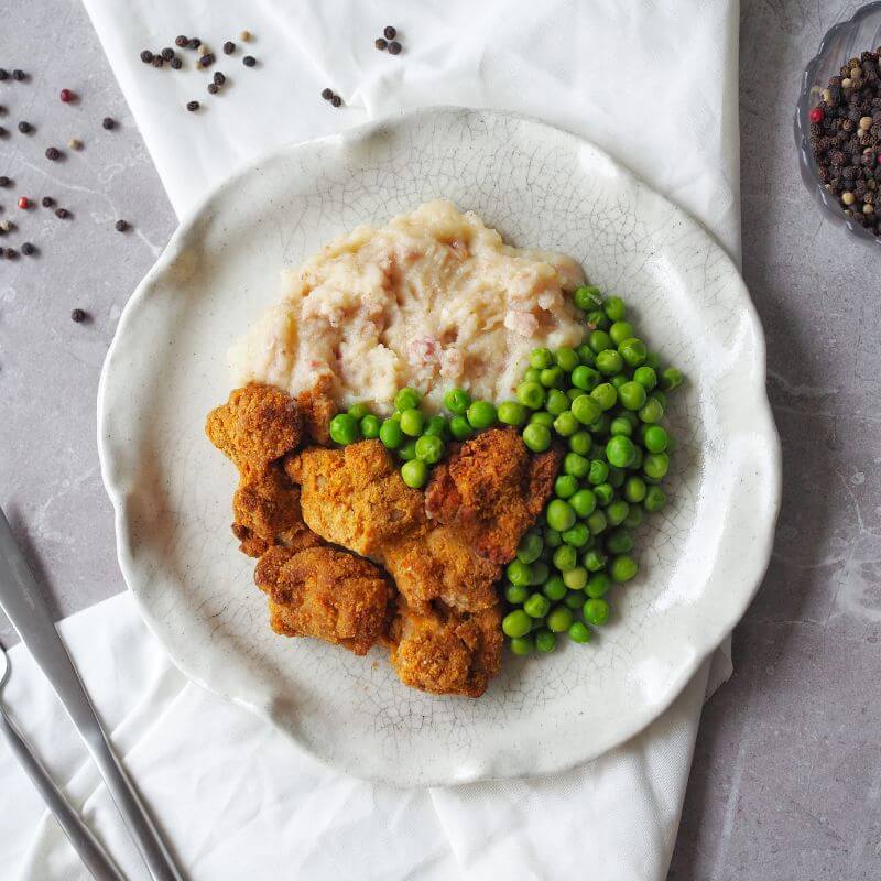 A plate of Popcorn Chicken with creamy cauliflower mash and peas, served on a white curved plate. The mash is made with cauliflower, bacon, and ghee, and the crispy popcorn chicken is coated in almond meal and spices. The dish is garnished with cracked peppercorns, scattered across a grey marble bench top. A white cloth is tucked beneath the plate, and a glass ramekin filled with cracked peppercorns is placed nearby. A fork and knife rest to the left of the plate.