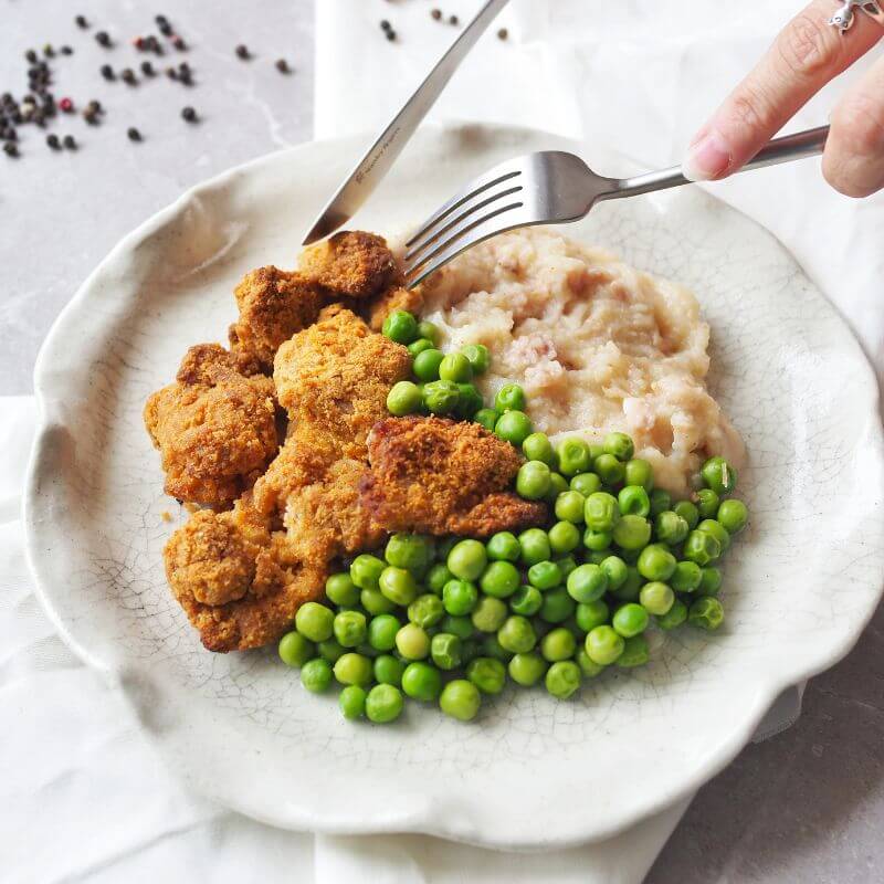 Two hands holding a fork and knife, cutting into the crispy popcorn chicken. The plate also features creamy cauliflower mash and peas. Cracked peppercorns are scattered on the white cloth and grey marble bench.