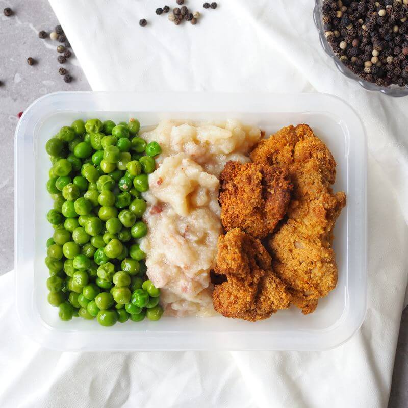 A clear meal container filled with Popcorn Chicken, creamy cauliflower mash, and peas. The mash is made with cauliflower, bacon, and ghee, while the chicken is seasoned and coated in almond meal. The white cloth is tucked underneath the container, and cracked peppercorns can be seen scattered nearby.