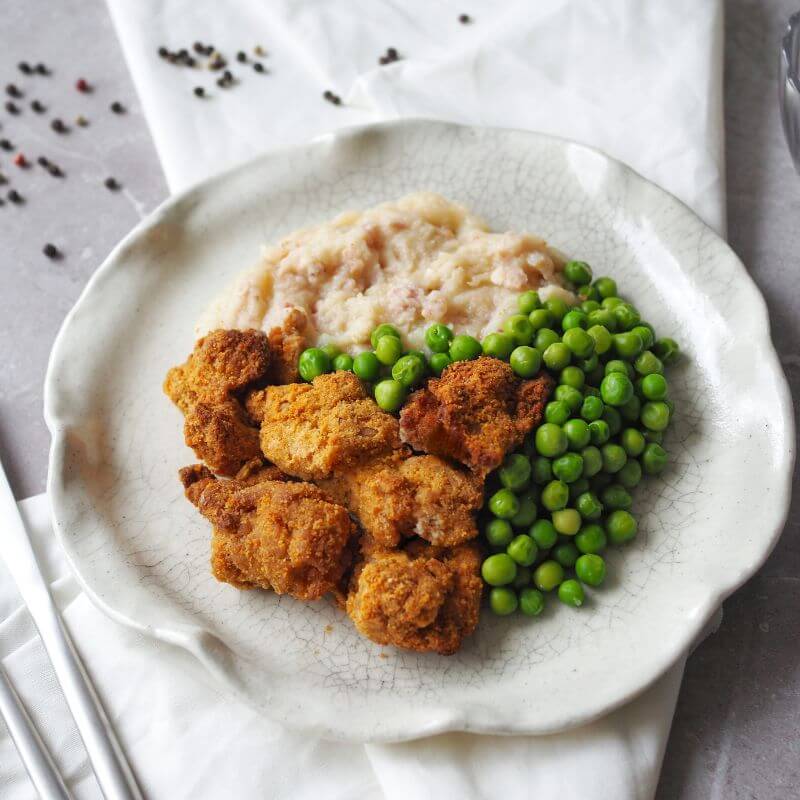 A close-up, side view of the Popcorn Chicken with creamy cauliflower mash and peas, served on a white curved plate. The dish highlights the crispy texture of the chicken and the smooth mash made with cauliflower, bacon, and ghee. Cracked peppercorns are scattered on the marble bench top, adding a touch of detail.