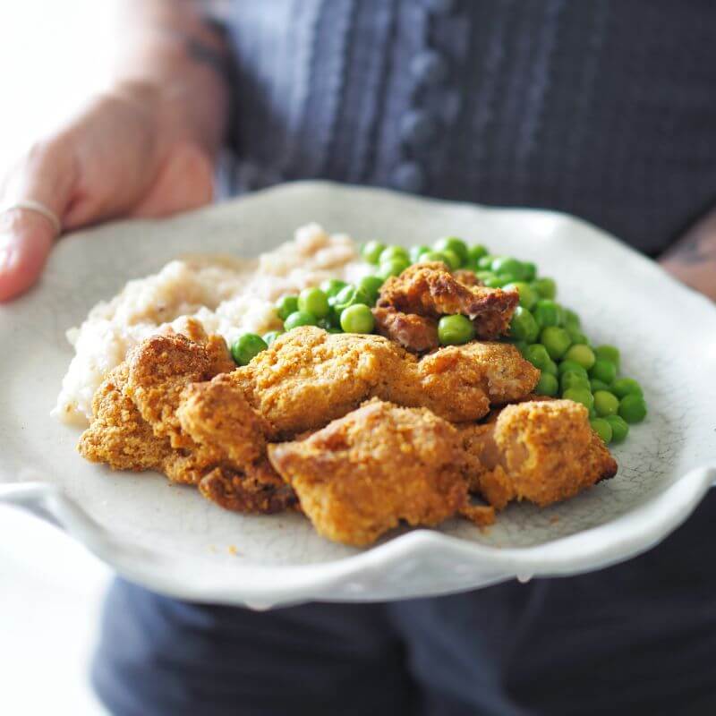 A woman wearing a navy-blue buttoned shirt holds a plate of Popcorn Chicken with creamy cauliflower mash and peas. The dish is displayed in a casual and inviting manner, emphasizing the golden, crispy coating of the chicken and the creamy mash.