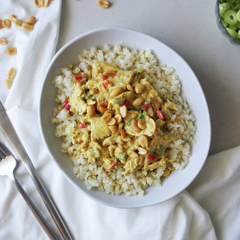 A white bowl filled with Satay Chicken served on a bed of fluffy cauliflower rice and vibrant capsicum. The tender chicken is coated in a rich peanut satay sauce and garnished with chopped spring onion. Scattered peanuts and a glass ramekin of chopped spring onion sit on the white cloth tucked beneath the bowl. A fork and knife are placed to the left of the bowl, completing the inviting presentation.