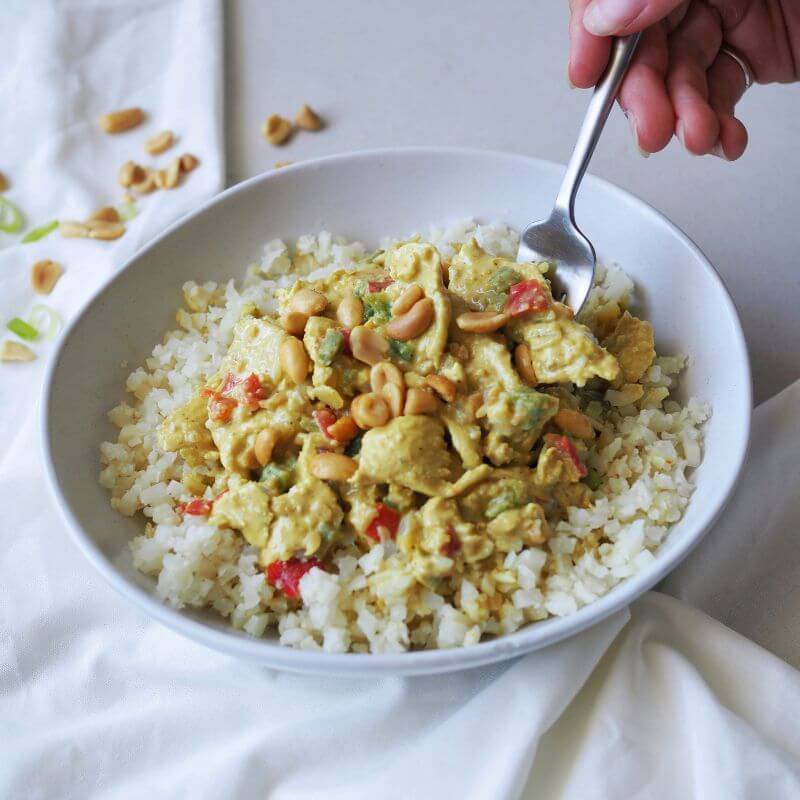 A hand holding a fork, cutting into the tender Satay Chicken coated in creamy peanut satay sauce. The dish sits on a bed of cauliflower rice with vibrant capsicum and is garnished with chopped spring onion. Scattered peanuts and a white cloth can be seen in the background.