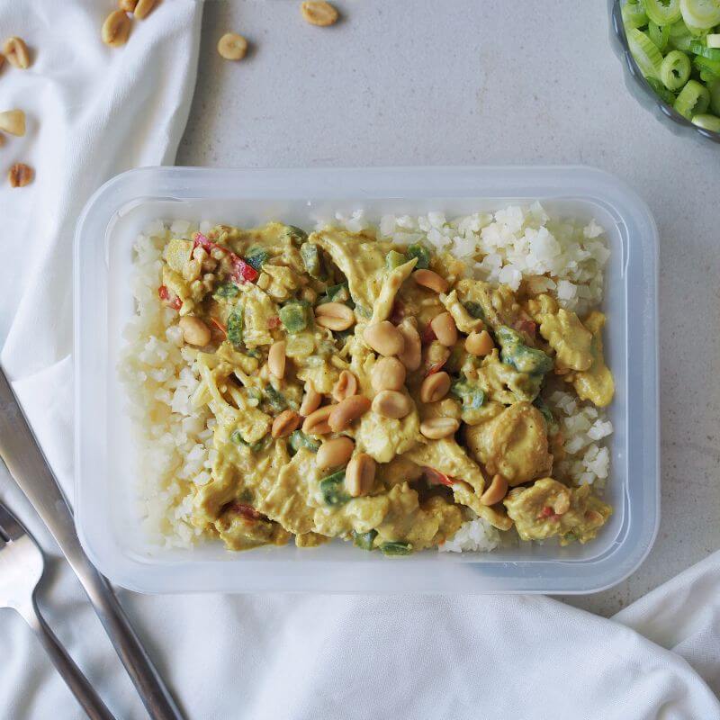 A clear meal container filled with Satay Chicken and cauliflower rice, accompanied by bright capsicum slices. The chicken is coated in a creamy peanut satay sauce and garnished with chopped spring onion. A white cloth is tucked beneath the container, with scattered peanuts nearby.