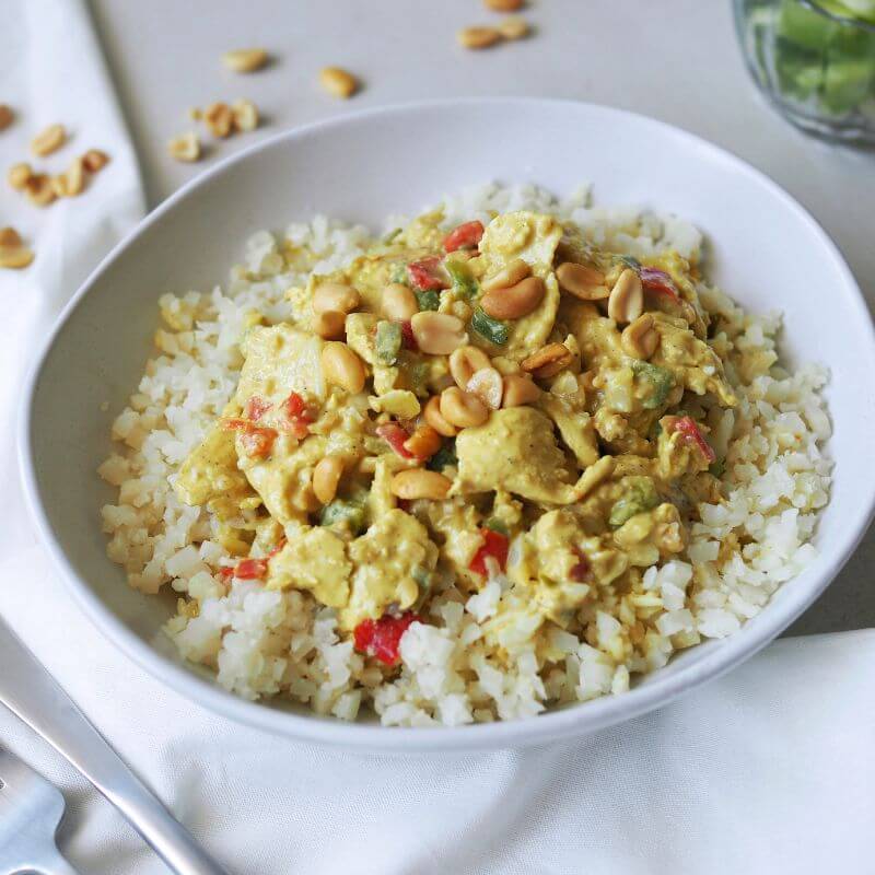 A side view of Satay Chicken served on a bed of cauliflower rice and capsicum. The creamy peanut sauce glistens over the tender chicken pieces, and the dish is garnished with fresh spring onion. Scattered peanuts add a rustic touch, while the white cloth and fork are visible in the background.