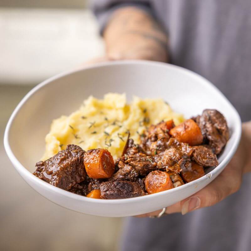 Two hands gently cradle a bowl of Beef Bourguignon, capturing the comforting and homely essence of this French-inspired dish with its deep, hearty flavors.