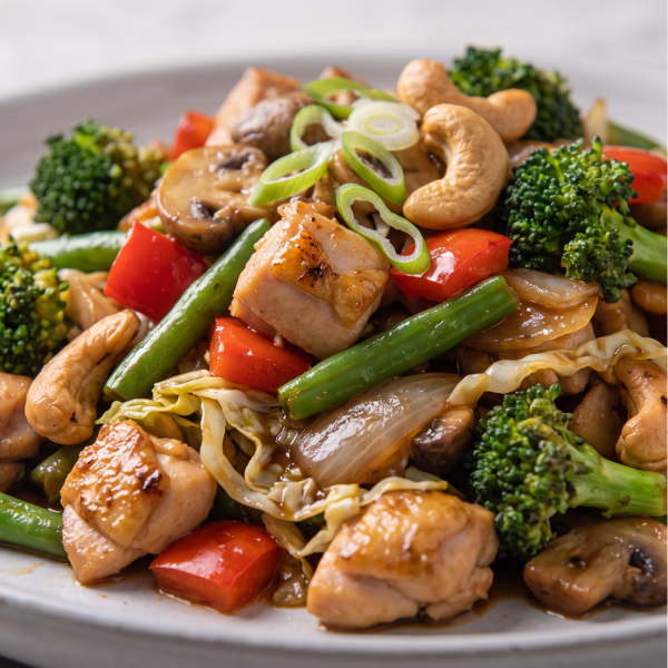 Close-up shot of stir-fried chicken, vibrant broccoli florets, green beans, sliced red capsicum, and whole roasted cashews coated in a glossy savoury sauce.