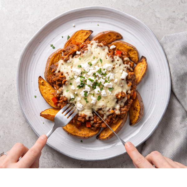 Overhead shot of loaded sweet potato wedged on a white ceramic plate. Hands are shown using knife and fork to cut into the delicious meal. 