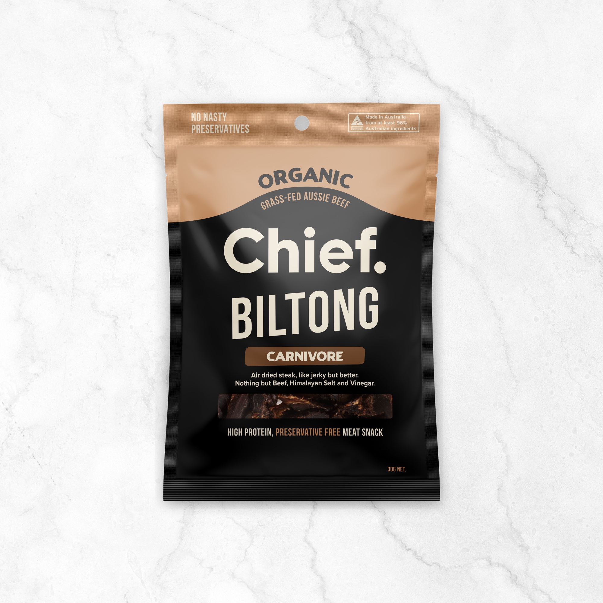 A black and tan 30g bag of Chief Carnivore Biltong standing upright on a white marble surface. The front reads “Organic grass-fed Aussie beef, Chief. Biltong, Carnivore”, with “High protein, preservative free meat snack” along the bottom.