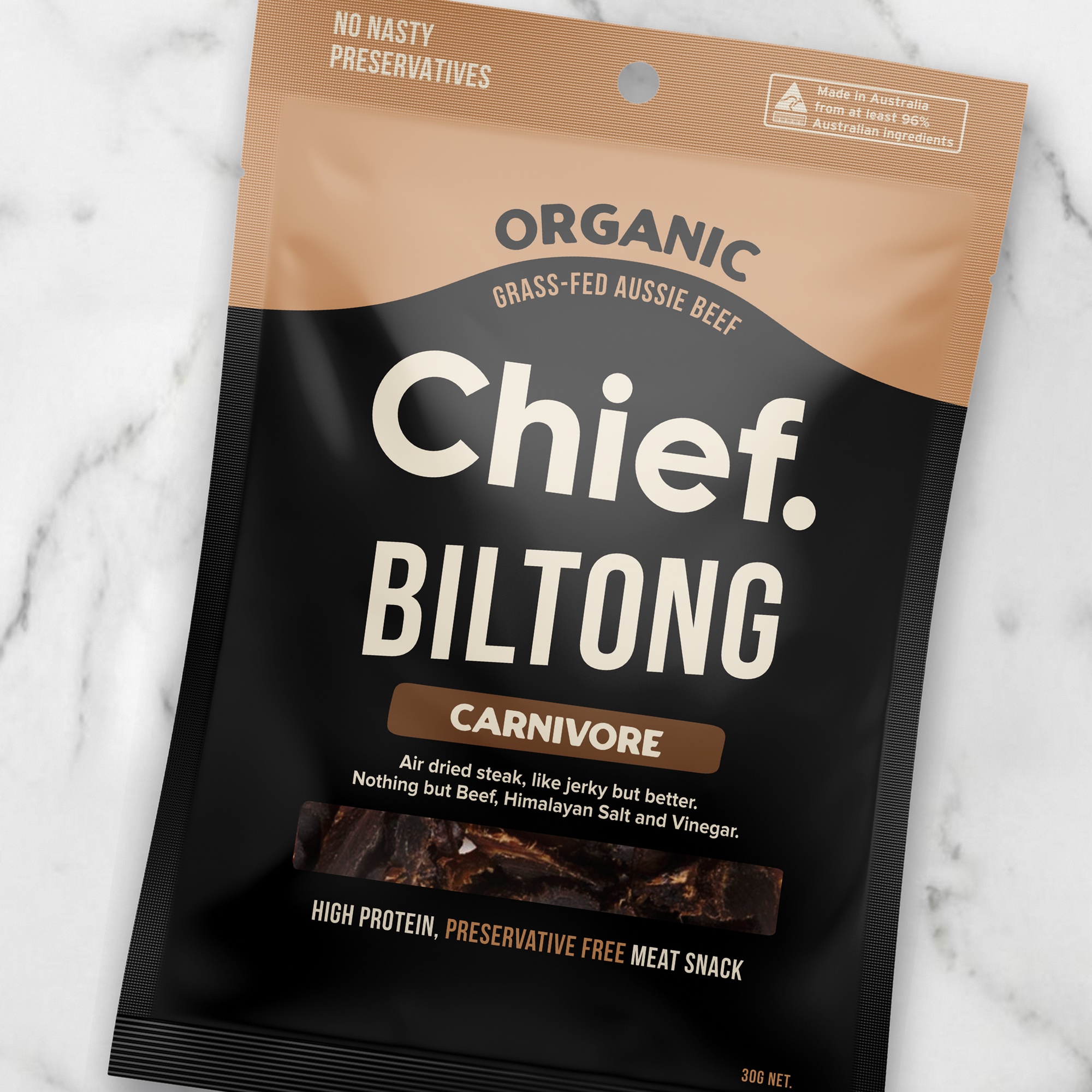 A close-up of the Chief Carnivore Biltong bag, tilted slightly on white marble. Large cream lettering reads “Chief. Biltong” above a tan “Carnivore” label. A line underneath says it’s air-dried steak, like jerky but better, made with nothing but beef, Himalayan salt and vinegar.