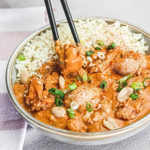 Action shot featuring black chopsticks picking up a piece of Kung Pao Chicken. The beige and white striped cloth adds a soft backdrop, emphasizing the sticky, rich texture of the low-carb peanut Kung Pao sauce.
